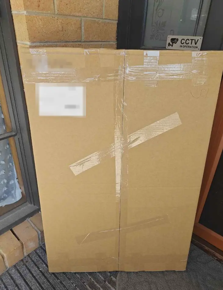 A flat cardboard carton of corflute signs, taped shut, standing against a front door beside a CCTV sign. The delivery label and job number are blurred.