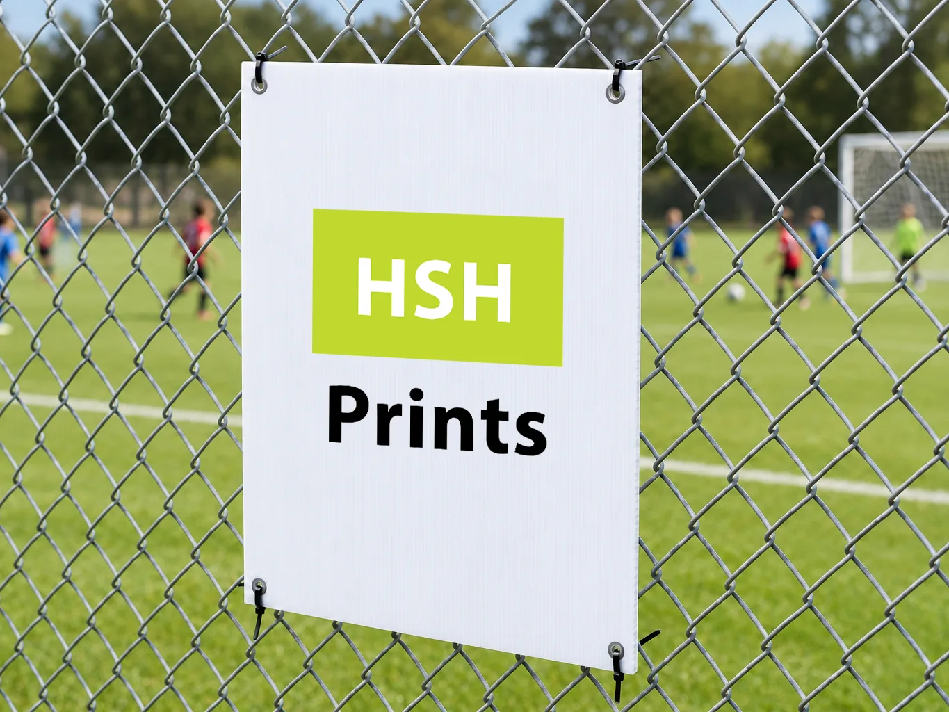 Mockup of a white corflute sign carrying the HSH logo, cable-tied by its corner eyelets to a chain-wire fence beside a sports field