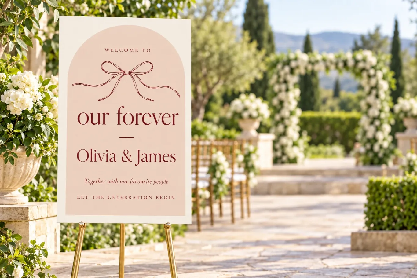 A blush-pink arched foam board wedding welcome sign, our forever, Olivia and James, on a gold easel in a garden