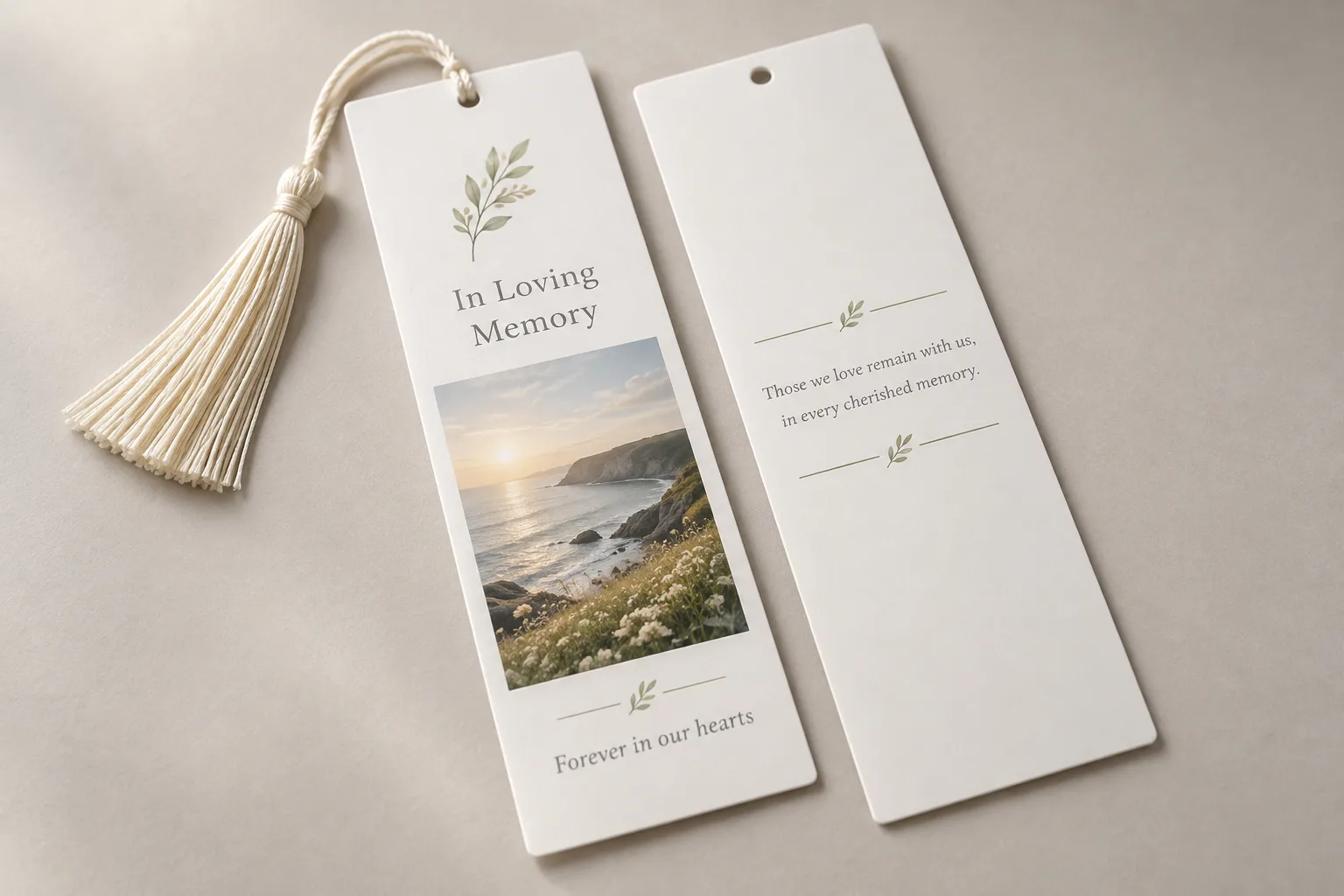 A memorial bookmark with a cream tassel, a leaf motif, In Loving Memory and a coastal photograph, beside its back with a short verse