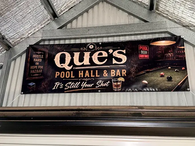 The printed vinyl banner reading Que's Pool Hall & Bar, It's Still Your Shot, hung across the gable of a steel shed.